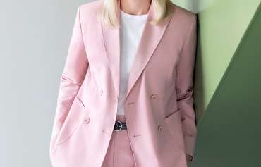 Nikola Steinbock (Sprecherin) Eine Frau mit glattem, schulterlangem, blondem Haar steht vor einer Wand mit geometrischen Flächen in Hellgrau und Pastellgrün. Sie trägt einen rosafarbenen Hosenanzug mit doppelreihigem Blazer und ein weißes T-Shirt. Ihre Hände sind locker in die Hosentaschen gesteckt. Sie blickt freundlich in die Kamera.